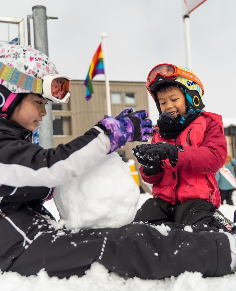 Two children sitting in the snow building a snowman.
