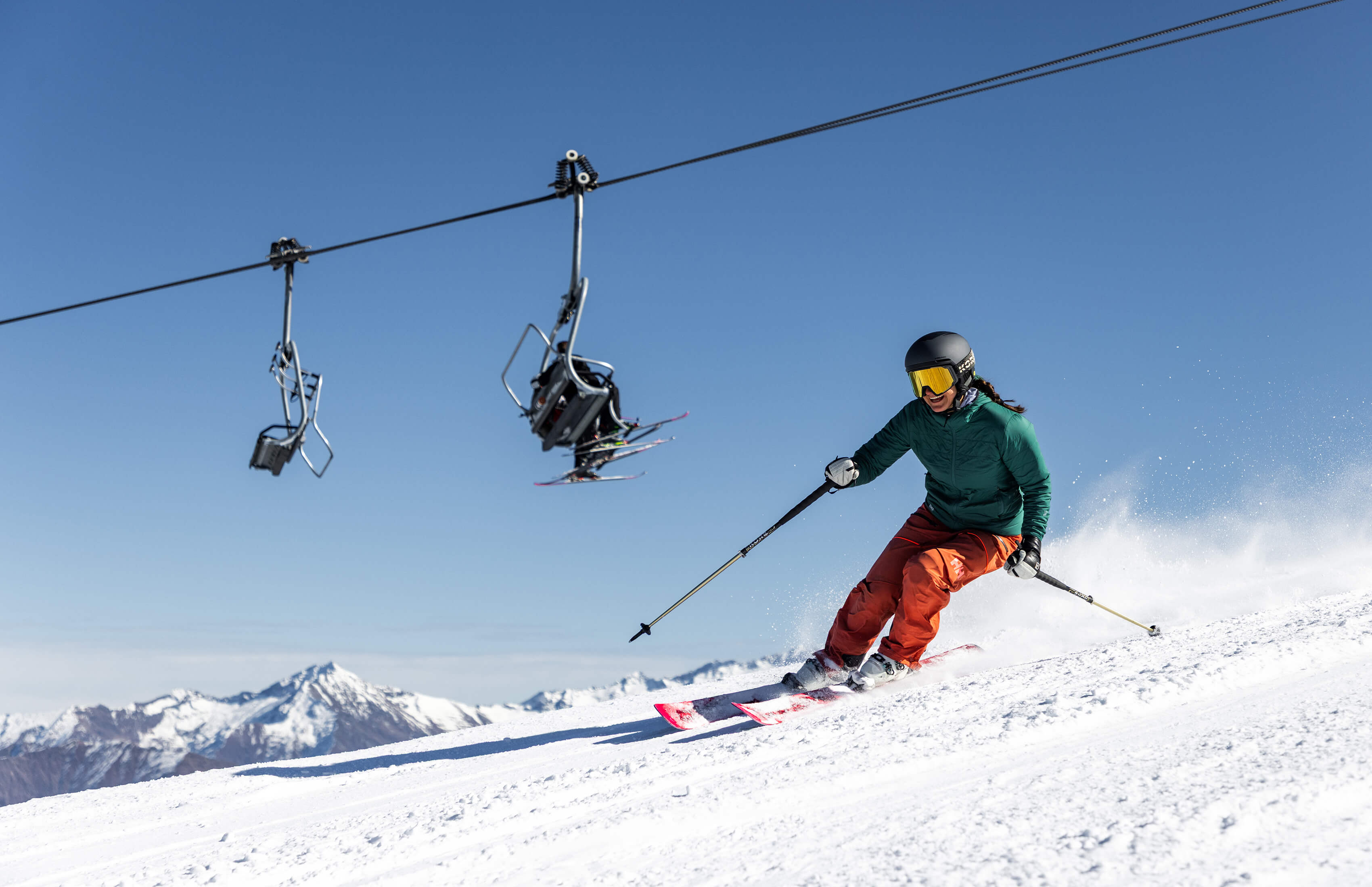 Skier in green jacket and orange pants skiing down snowy slope with lift chairs and mountains behind.