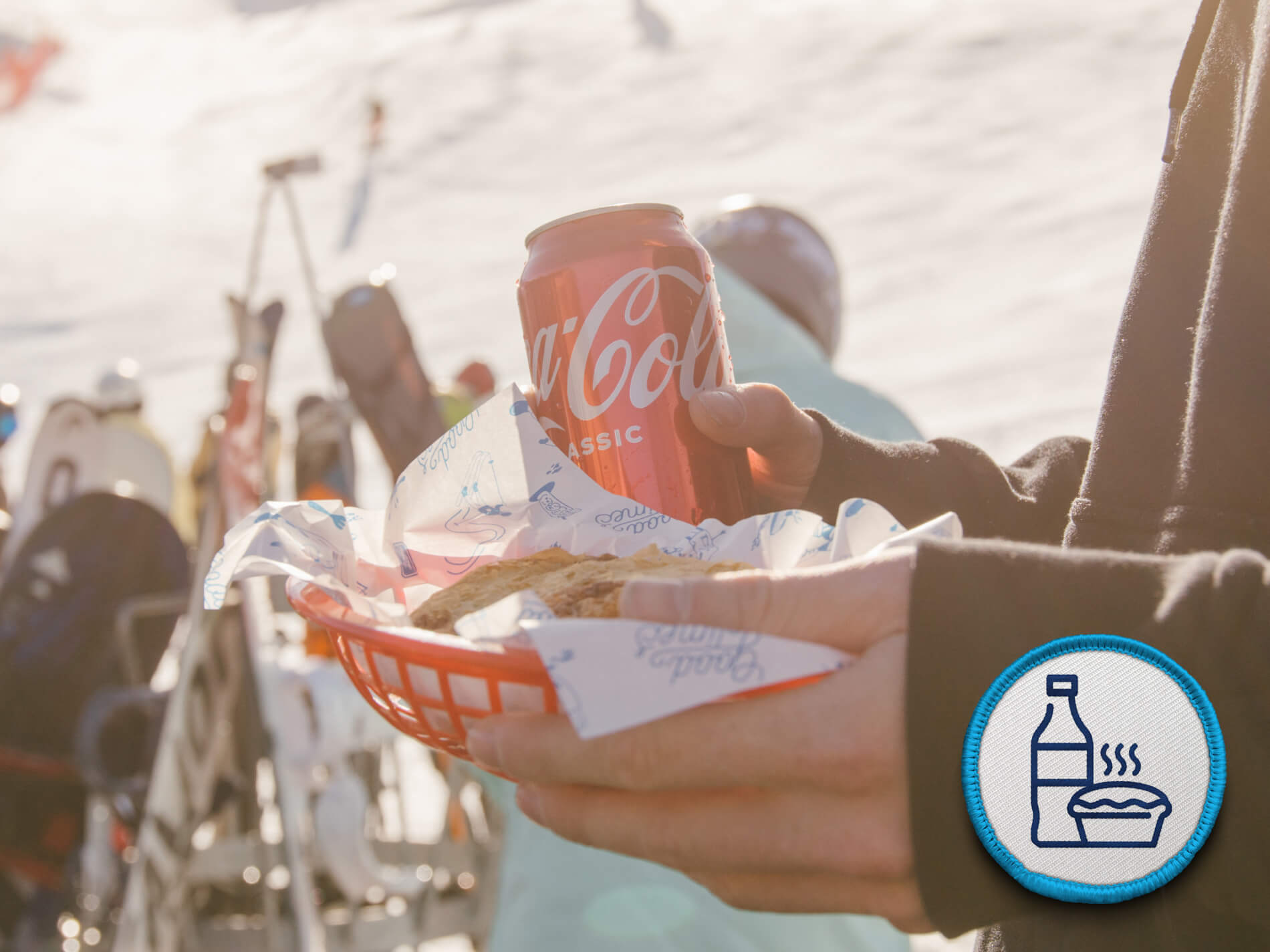 Person holding a basket with a can of Coca-Cola Classic and a wrapped food item outdoors, skis in background.