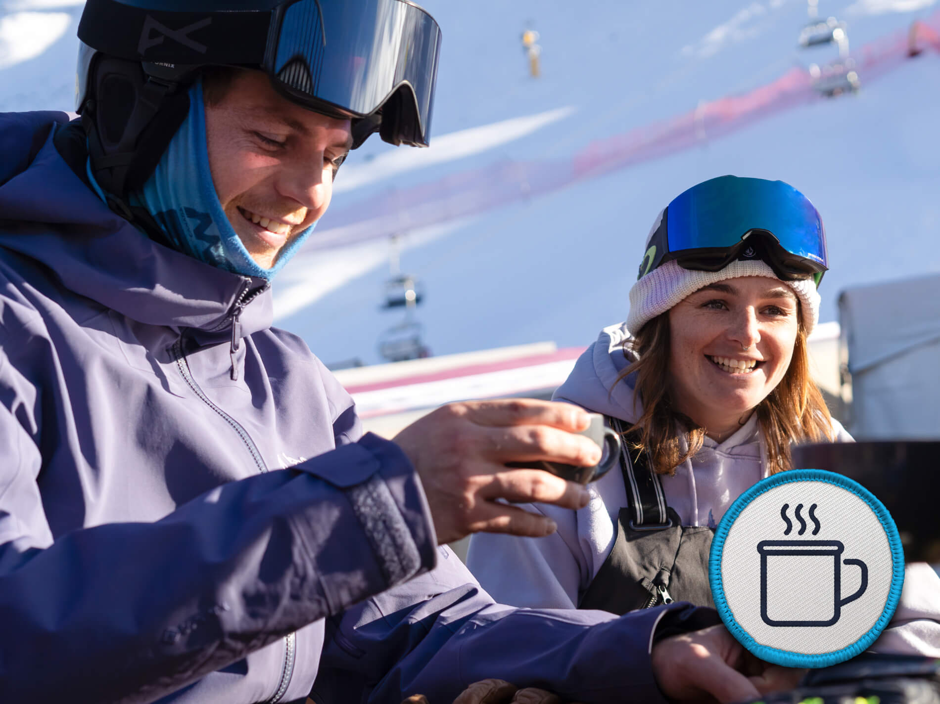 Two skiers outdoors smiling and holding a warm drink with snowy mountains behind them.