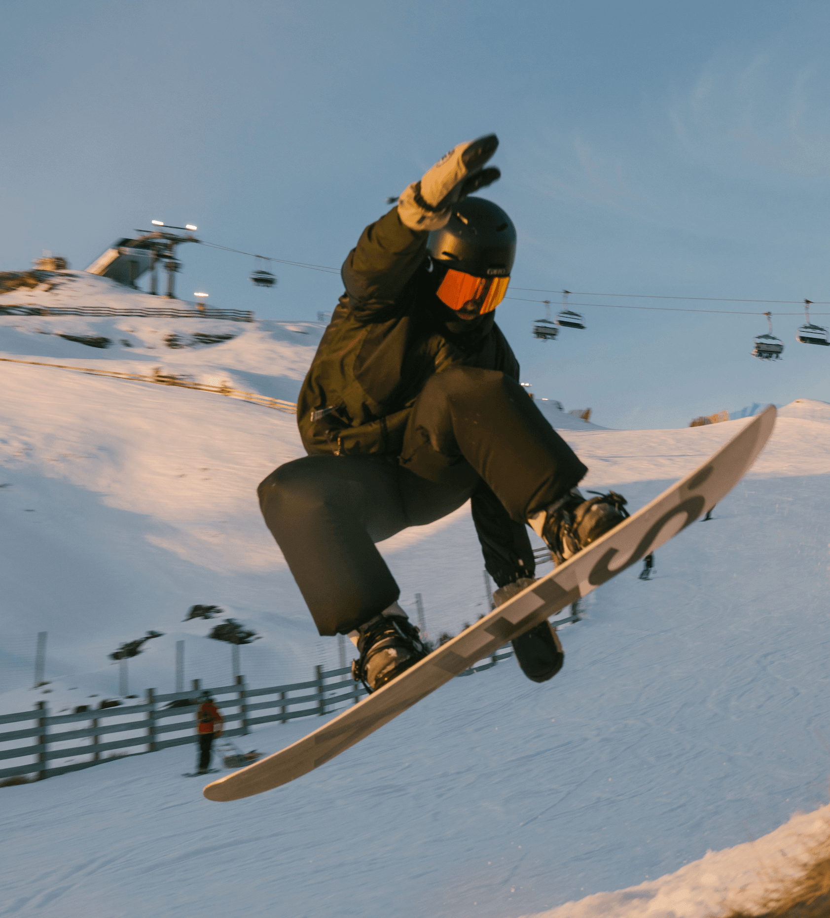 Snowboarder in black gear performs a jump on snowy slope with ski lifts in the background.
