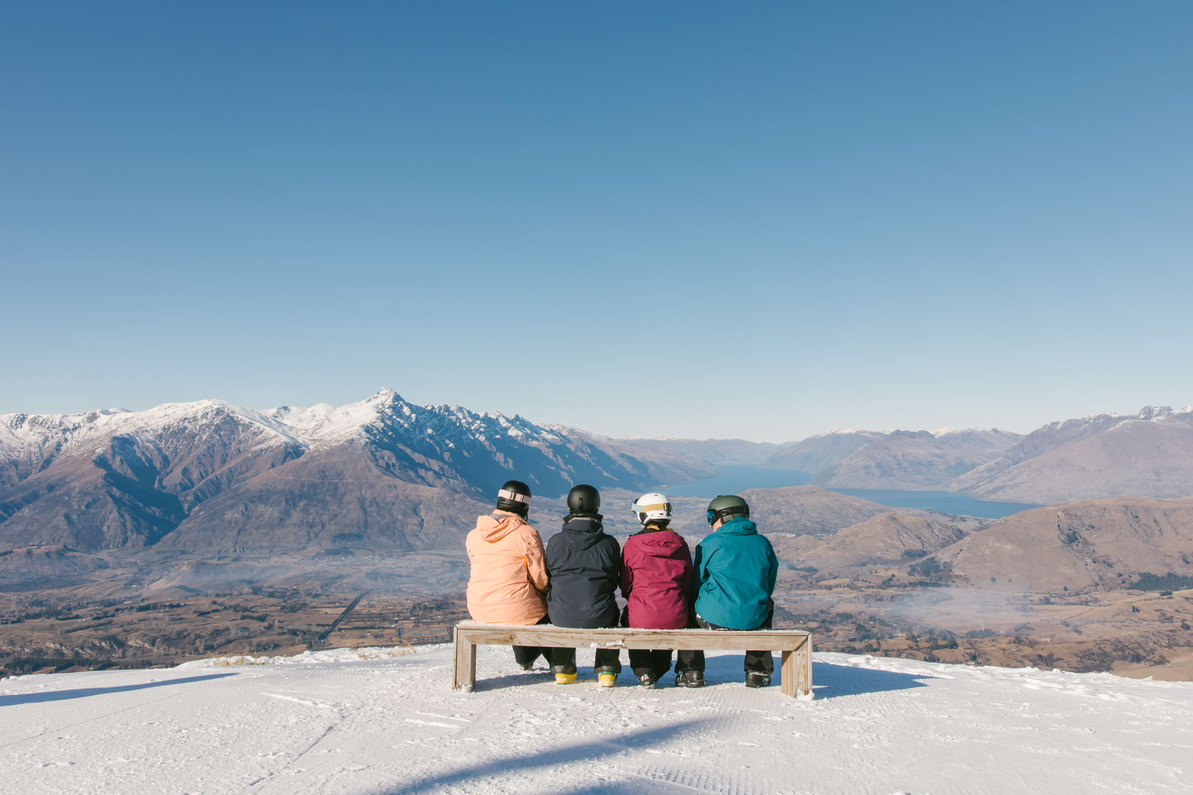 Four people in winter gear sitting on a bench facing snowy mountains and a blue lake under clear sky.