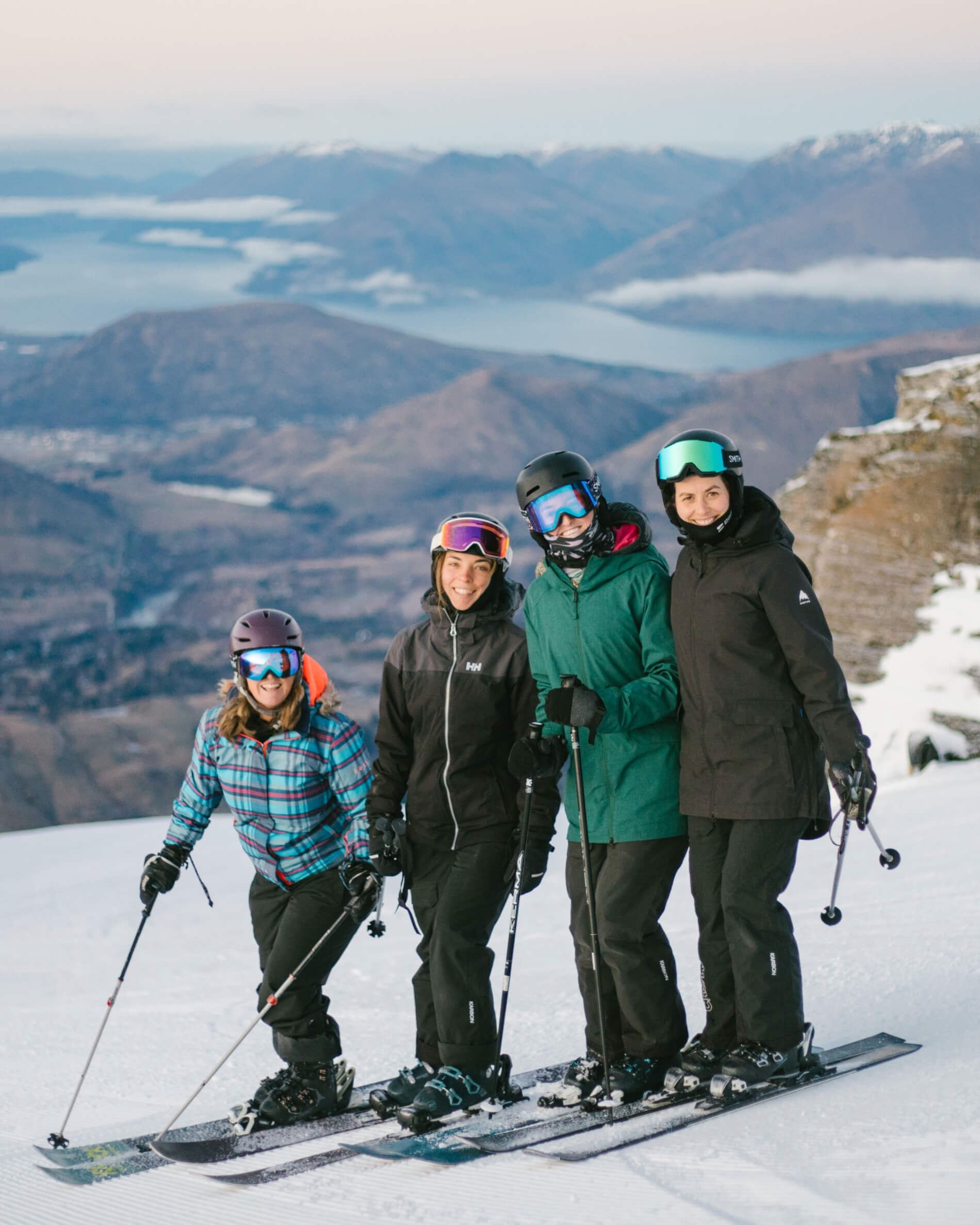 Four skiers smiling at camera on snowy slope with mountains and lake in background.
