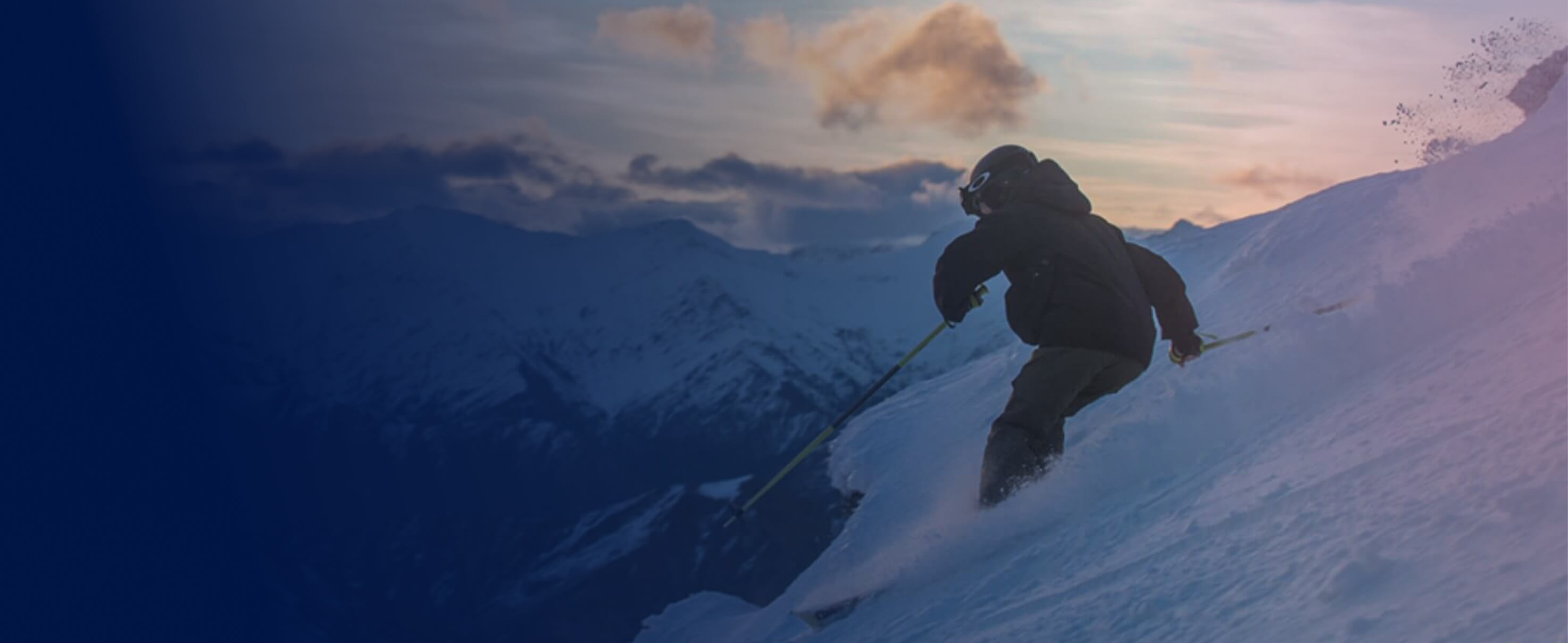 Skier in black jacket skiing down a snowy mountain slope at sunset with mountains in background.
