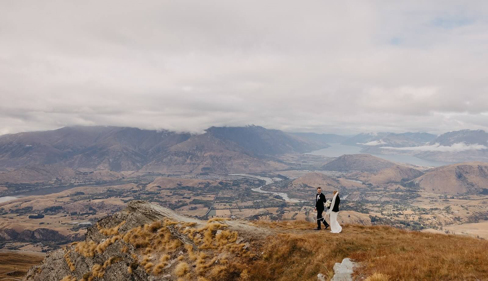 Bride and groom walking on grassy hilltop with a vast mountainous valley and river behind them.