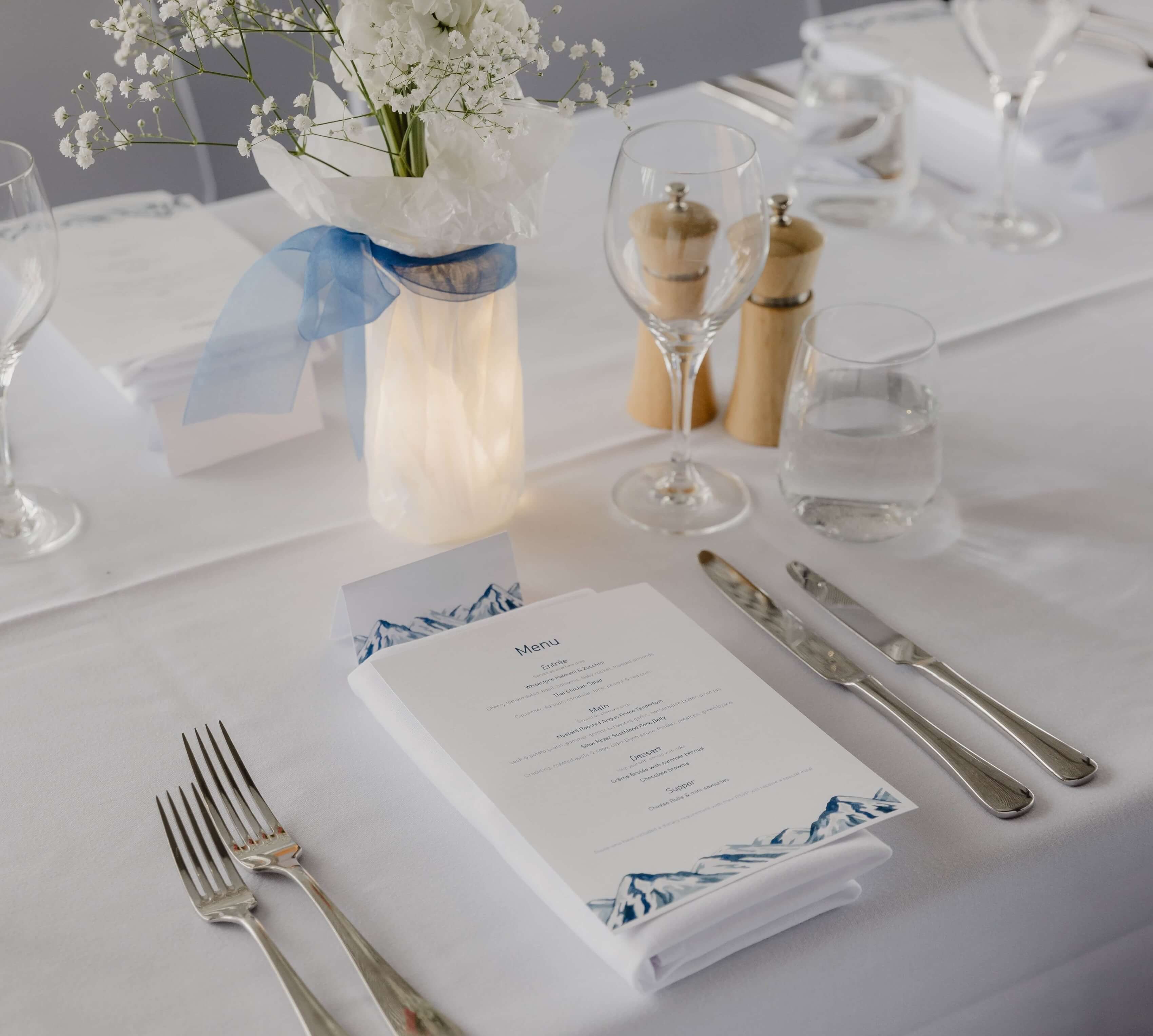 Elegant white table setting with menu, silverware, glassware, flowers in vase, and blue ribbon.