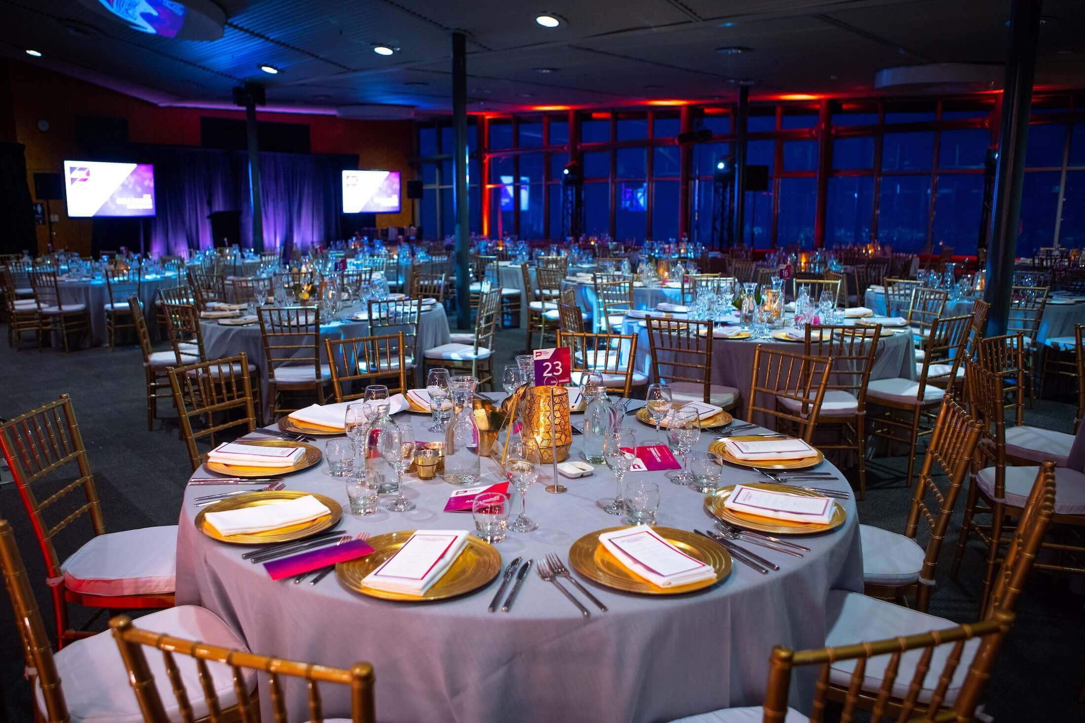 Round event tables with gold plates and glassware in a dim venue lit with blue and red lights.