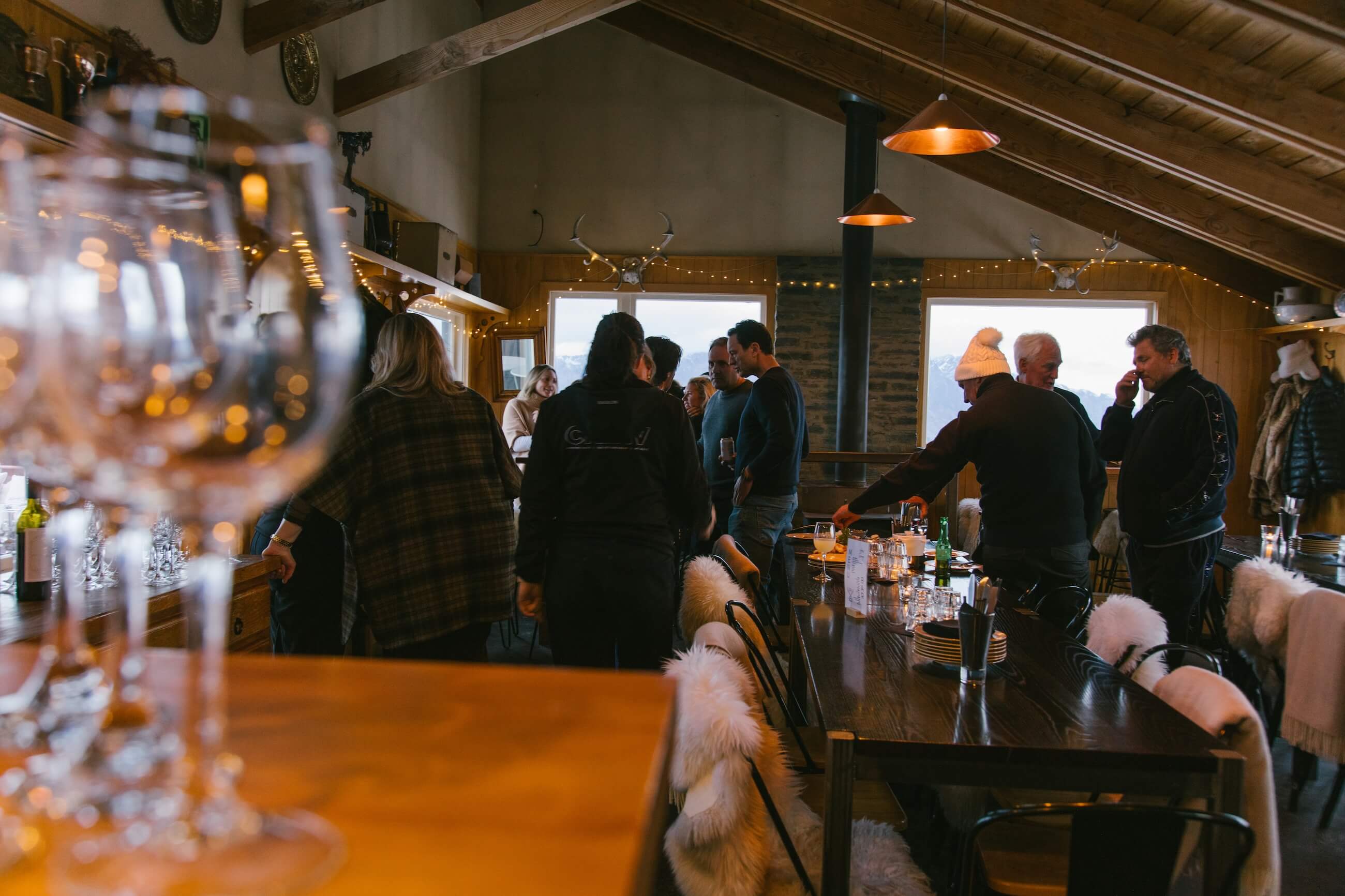 Group of people socializing inside a cozy wooden cabin with tables, chairs, and hanging lamps.