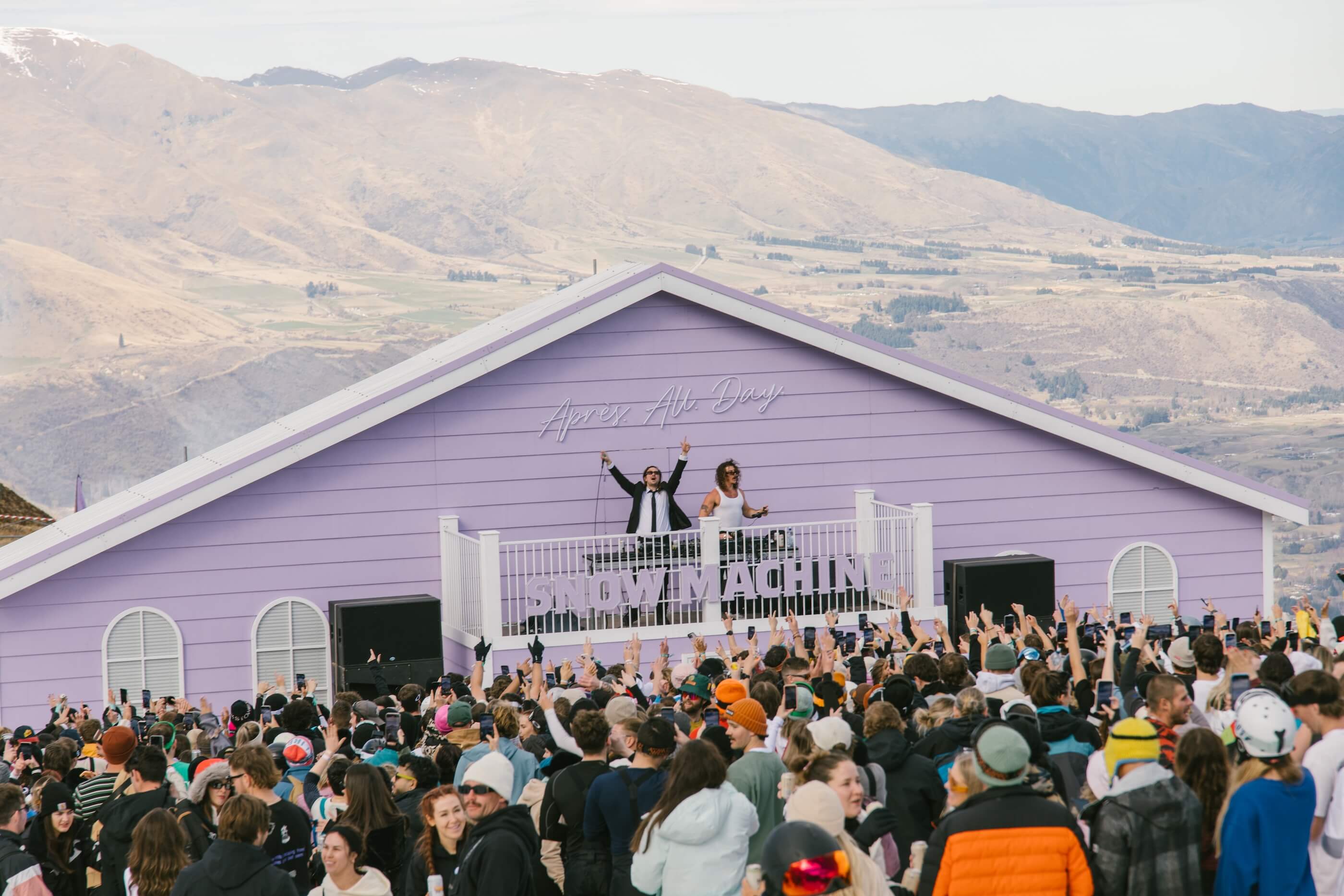 Large crowd watches two DJs perform on a purple stage labeled Snow Machine with mountains behind.