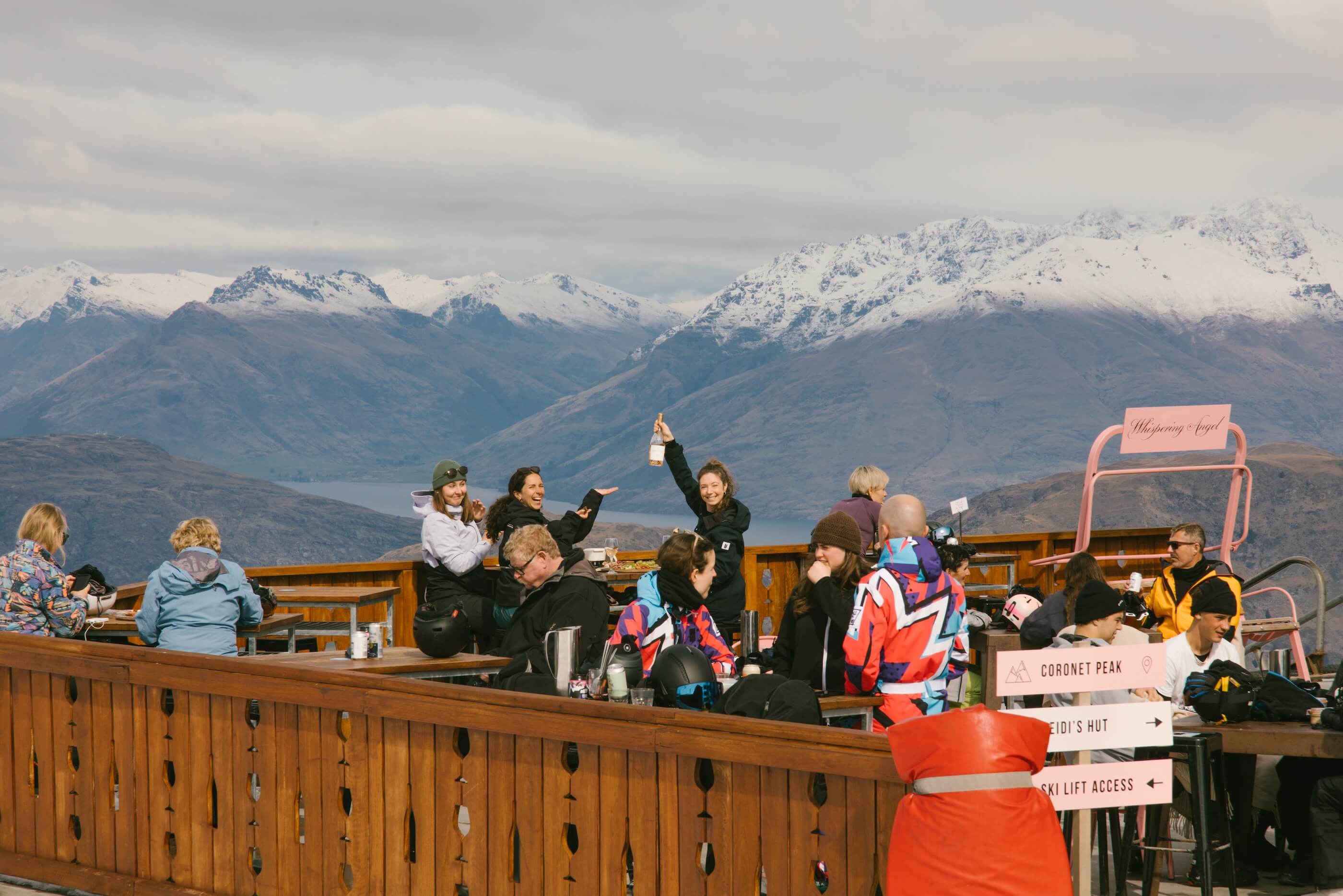 Group of people enjoying drinks on outdoor wooden deck with snow-capped mountains in the background.