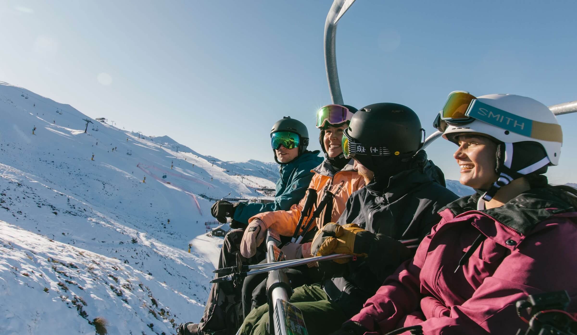 Four skiers wearing helmets and goggles riding a ski lift over snowy mountain slopes on a clear day.