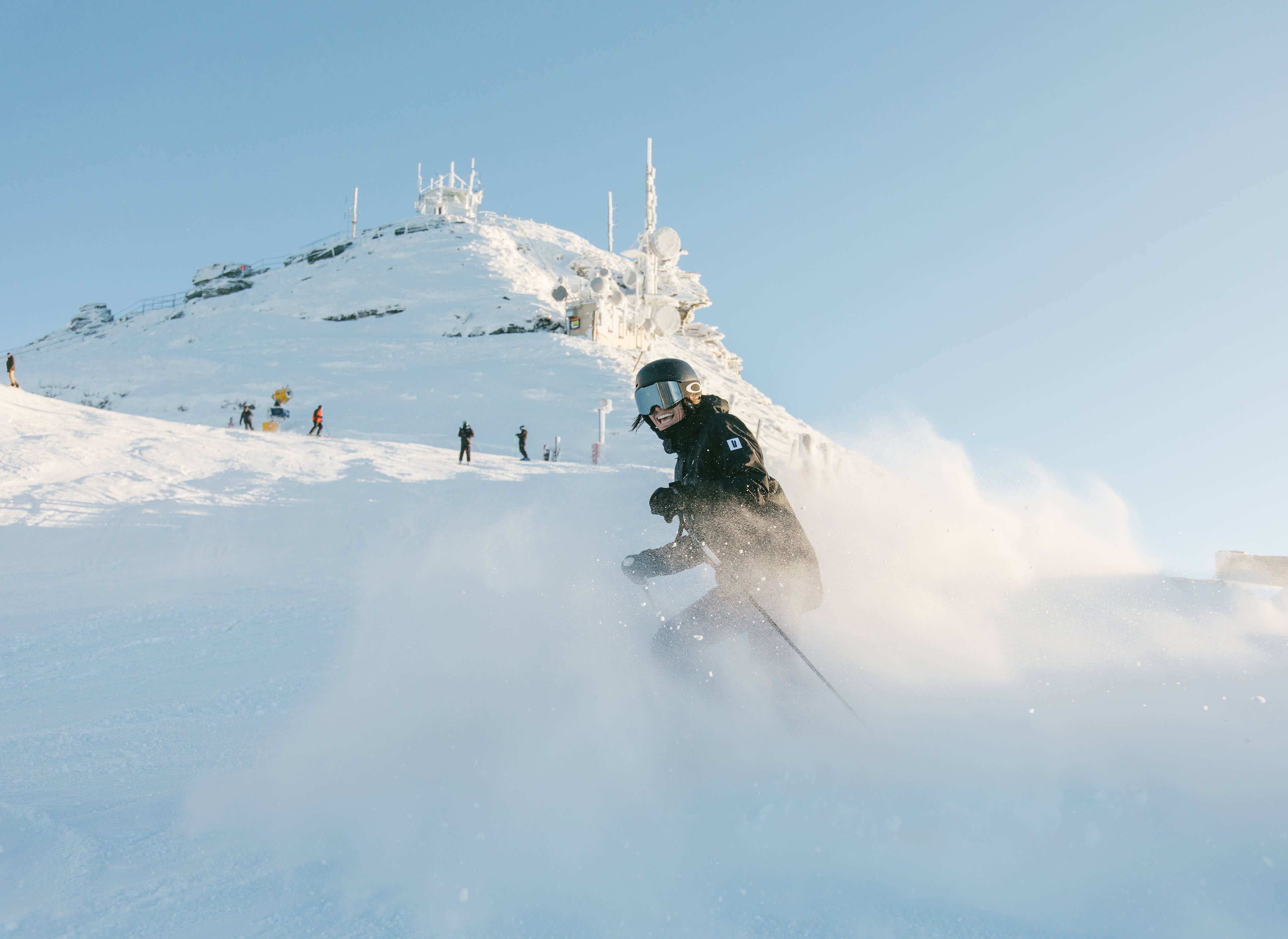 Smiling skier in black gear skiing down a snowy mountain with others and antennas in background.