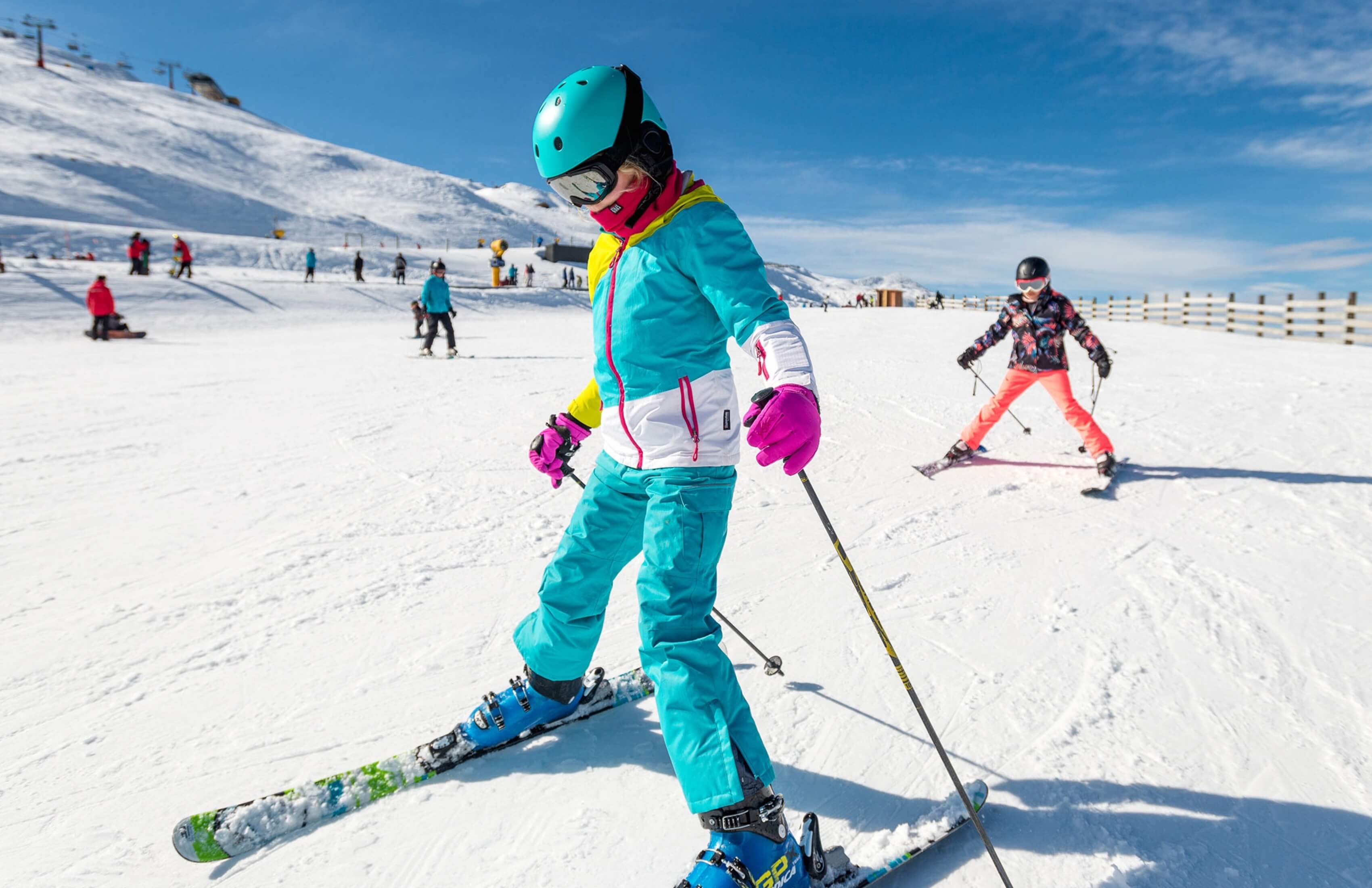 Two children skiing on a snowy slope under a clear blue sky with ski lifts in the background.