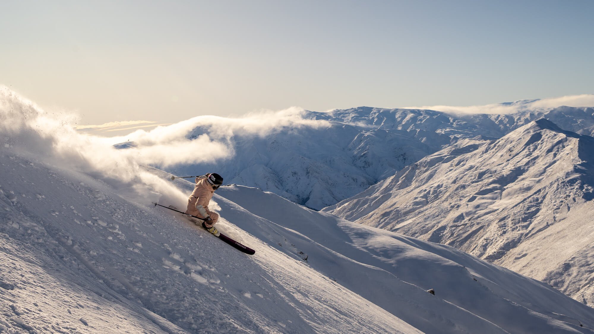 Skier in tan outfit carving down a snowy mountain slope with snow-covered peaks in the background.