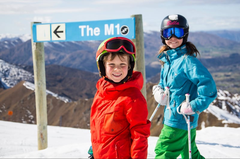 Two children smiling in front of a ski trail sign labelled 'The M1' at Coronet Peak
